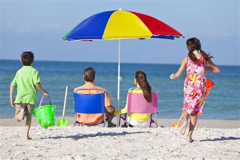 Four people at the beach, two adults sitting in chairs under a multicoloured umbrella looking out to sea and two children running away from us towards them
